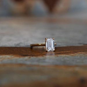 Könnte beinhalten: Ein goldener Verlobungsring mit einem Diamanten im Smaragdschliff. Der Ring hat ein schlankes, poliertes Band, und der Diamant ist mit vier Krappen gefasst. Der Diamant ist klar und reflektiert das Licht.
