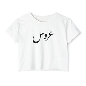 May include: White crop top with a crew neck and short sleeves. The shirt features black Arabic script across the front. The fabric appears to be a soft, lightweight material. The overall style is casual and modern.