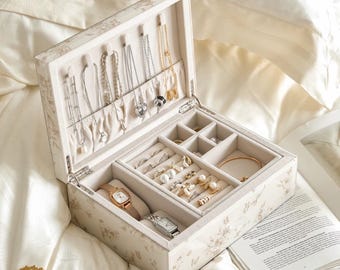 Può includere: Portagioie aperto con motivo floreale color crema e beige. L'interno è foderato in tessuto beige chiaro e contiene collane, anelli, orecchini e due orologi. Scatola per riporre gioielli.