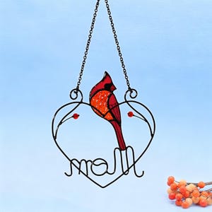 May include: A stained glass cardinal bird ornament with a black metal heart-shaped frame. The cardinal is red and orange, with a black head. The word "Mom" is written in cursive at the bottom of the heart. The ornament hangs from a chain.