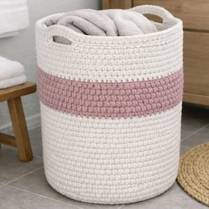 Może przedstawiać: Cylindryczny kosz na pranie ze strukturalnym pasem. Kosz wykonany jest z grubej, kremowej przędzy z teksturowanym, jasnoróżowym pasem pośrodku. Kosz ma dwa uchwyty i jest wypełniony złożonymi ręcznikami.