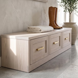 May include: A light-toned wooden storage bench featuring three drawers with rectangular recessed panels and horizontal brushed gold metal pulls. A folded cream-colored textile rests on the flat top surface next to a pair of tall tan leather boots.