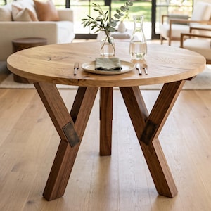 May include: A round wooden dining table with a natural grain finish, featuring three angled legs connected by dark metal plates. On the tabletop sits a ceramic plate with a folded green napkin, a fork, a knife, a spoon, a glass vase with greenery, and a glass carafe.