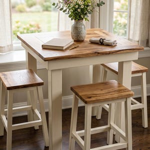 May include: A square wooden table with a natural wood grain top and cream-colored legs, accompanied by three matching stools. On the table sits a ceramic vase filled with wildflowers, a closed beige book, a gold pen, and a folded napkin.