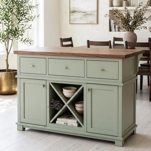 May include: A sage green kitchen island featuring a rectangular dark wood countertop, three top drawers with small gold knobs, two side cabinets, and a central X-shaped open shelving unit containing two ceramic bowls and two stacked books.