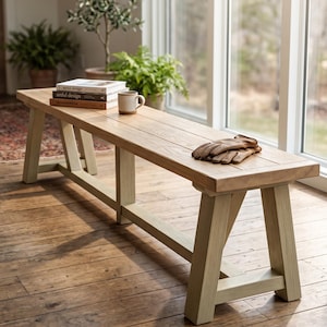 May include: A long wooden bench with a natural light-toned wood grain top and pale green painted legs. The bench features a sturdy trestle-style base with cross-bracing. A stack of three books and a ceramic mug sit on one end, with leather gloves.