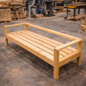 Peut inclure: Un banc en bois clair avec une assise à lattes et des accoudoirs. Le banc est rectangulaire et semble être nouvellement construit. Le bois a une finition naturelle, mettant en valeur le grain. Le banc est dans un atelier.
