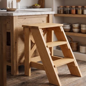 May include: A light-colored wooden step stool featuring three tiers, including a top platform and two lower steps. The structure is supported by angled side panels with a cross-brace design, providing a stable base for reaching high areas.
