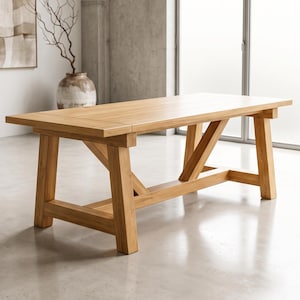May include: A rectangular light brown wooden dining table featuring a smooth flat top surface and a sturdy trestle base with angled support beams connecting the legs to a horizontal floor stretcher bar, showcasing a natural wood grain finish.