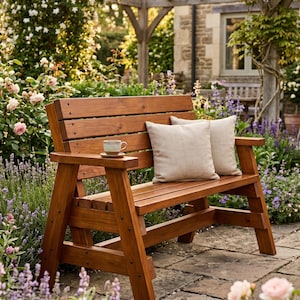 May include: A wooden outdoor bench with a slatted back and armrests. Two beige throw pillows rest on the seat. A small teacup and saucer sit on the armrest. The bench is surrounded by a garden with flowers and greenery.