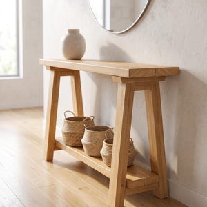 May include: A light-toned wooden console table with a rectangular top and a lower shelf. The table features angled legs and a sturdy frame. Three woven seagrass baskets sit on the lower shelf, and a single off-white ceramic vase rests on top.
