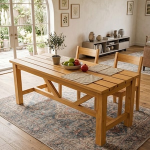Puede incluir: Una mesa de comedor de madera marrón claro con dos sillas a juego. La mesa tiene una parte superior de listones y una base de travesaños. Un cuenco de fruta y una pequeña planta en maceta están sobre la mesa. La mesa está sobre una alfombra estampada.