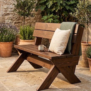 May include: A dark brown wooden bench with an X-frame design. A beige pillow and a green blanket rest on the bench. A small mug sits on the bench. Potted plants and a stone wall are in the background.