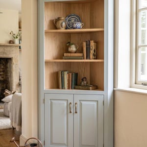 May include: A tall corner cabinet featuring a light blue painted exterior frame and natural wood interior shelving. The unit includes two lower cabinet doors with brass-toned handles and three open shelves displaying various ceramic items and books.