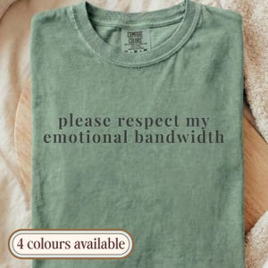 Puede incluir: Camiseta verde salvia con la frase "please respect my emotional bandwidth" impresa en gris oscuro. La camiseta tiene cuello redondo y una etiqueta que dice "Comfort Colors". La imagen también incluye un pequeño texto que dice "4 colores disponibles".