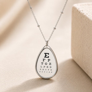 Op de afbeelding: Zilverkleurige ketting met een druppelvormige hanger met een klassieke oogtestkaart. De hanger toont zwarte letters op een witte achtergrond, met een fijne ketting en kleine kraalaccenten.