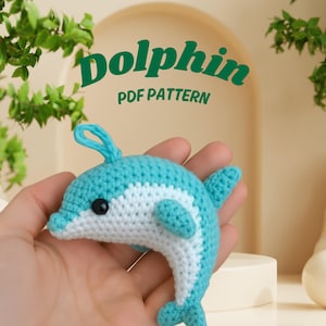 Könnte beinhalten: Ein gehäkelter Delfin in Blau- und Weißtönen mit einem schwarzen Auge. Der Delfin wird in einer Hand gehalten. Der Text "Dolphin PDF PATTERN" wird grün über dem Delfin angezeigt.