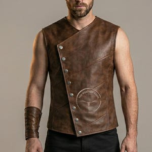May include: Brown leather vest with a unique asymmetrical button closure. The vest features a round logo on the lower right side and is sleeveless. The vest is a medium brown color and has a rugged, textured appearance.