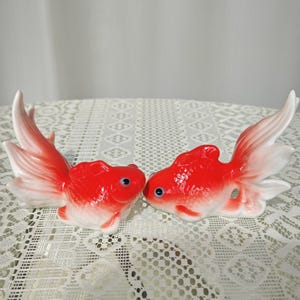 Könnte beinhalten: Zwei Keramik-Goldfischfiguren. Die Fische sind rot mit weißen Akzenten an Flossen und Schwänzen. Sie haben blaue Augen und sind einander zugewandt. Die Figuren stehen auf einer weißen Spitzendecke.