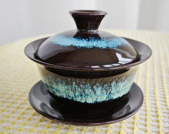 Puede incluir: Un juego de té gaiwan de cerámica marrón oscuro que consta de un cuenco, una tapa y un platillo. El exterior del cuenco y la parte superior de la tapa presentan un esmalte reactivo con un patrón de goteo en tonos turquesa y azul claro.
