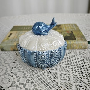 Puede incluir: Caja de baratijas de cerámica con un diseño azul y blanco. La tapa está rematada con una figura de ballena azul. La caja tiene un patrón texturizado, similar a un erizo de mar. La base es azul oscuro con un patrón circular.