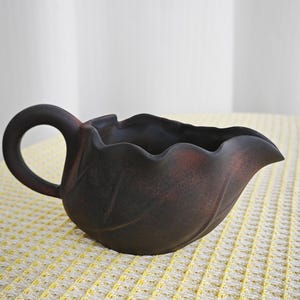 May include: A dark brown ceramic creamer or gravy boat shaped like a leaf with a curved handle and a scalloped rim. The surface has a matte, textured finish with subtle reddish-brown highlights along the edges and embossed leaf vein details.
