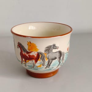 Puede incluir: Una taza de cerámica de color crema con una escena pintada de tres caballos en tonos marrón, amarillo y gris. La taza tiene un cuerpo redondeado, un borde acampanado y una base, todos acentuados con un borde de color marrón rojizo.