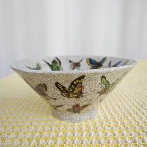 May include: A decorative ceramic bowl with a cream-colored, crackled finish. The bowl is adorned with colorful butterfly illustrations in shades of yellow, orange, blue, and green. The bowl has a flared shape and a small, round base.
