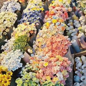 May include: A display of colorful ceramic flowers and plants in various pots. The flowers include roses, daisies, and other blooms in shades of yellow, pink, blue, and white. Some pots contain green leafy plants.