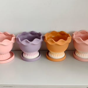 Puede incluir: Cuatro macetas de cerámica con bordes ondulados y platos a juego. Las macetas presentan un diseño degradado, pasando de colores pastel sólidos—rosa claro, lavanda, naranja y melocotón—en la parte superior a una base blanca en la parte inferior.