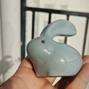Op de afbeelding: Lichtblauwe keramische konijnenfiguur met een craquelé-afwerking. Het konijn heeft een klein bruin stipje als oog. Het decoratieve item wordt in een hand gehouden.