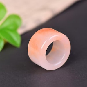 May include: A chunky ring with a cylindrical design. The ring is predominantly pale pink, with orange streaks and a polished surface. The ring is made of a translucent material, possibly jade or a similar stone.