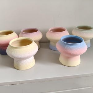 May include: Six mushroom-shaped ceramic planters with a speckled finish. Each planter features a unique pastel gradient colour scheme, including combinations of pink, yellow, blue, and purple. The pots have a wide, rounded top and a narrow base.