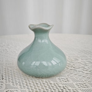 Pode incluir: Um pequeno vaso de cerâmica verde-claro com um corpo bulboso e uma borda ondulada. O vaso tem um esmalte craquelado e está sobre uma toalha de mesa de renda branca. O vaso tem cerca de 15 cm de altura.