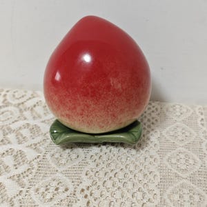 May include: A ceramic peach figurine with a glossy red finish, resting on a green leaf-shaped base. The peach has a gradient of red to light yellow. The figurine is a decorative item.