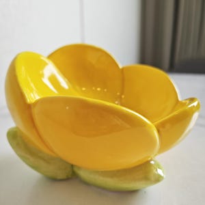May include: A ceramic bowl in the shape of a yellow flower with green detailing. The bowl has a glossy finish and petal-shaped edges. The bowl is likely for decorative use or to hold small items. The bowl's dimensions are approximately 15 cm in diameter.