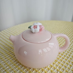 May include: A light pink ceramic teapot with a rounded body, a curved handle, and a short spout. The lid features a decorative sculpted floral knob with white and soft pink petals and a small green leaf accent, adding detail to the smooth finish.