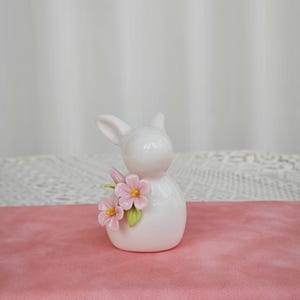 Op de afbeelding: Een wit keramiek konijnbeeldje met roze bloemen en gele centra. Het konijn heeft lange oren en is versierd met twee roze bloemen en groene bladeren. Het beeldje staat op een roze ondergrond.