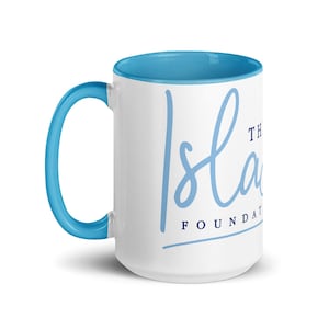 Puede incluir: Taza de cerámica blanca con interior y asa azul claro. La taza presenta el texto "Isla" en una fuente de escritura azul claro, con "THE FOUNDATION" debajo en una fuente más pequeña sin serifa.