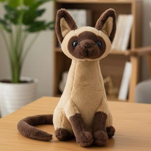 Può includere: Un peluche a forma di gatto siamese con corpo beige, muso, orecchie e zampe marroni scuro e occhi blu. Il gatto è seduto con la coda arricciata. Il giocattolo è su un tavolo di legno.
