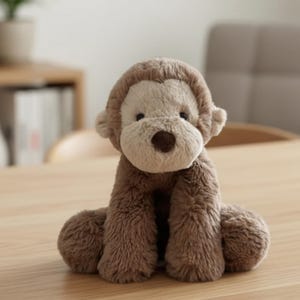 May include: A plush monkey toy with brown fur and a light tan face, ears, and hands. The stuffed animal is sitting on a wooden surface. The monkey has a round brown nose and small black eyes.