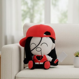 Puede incluir: Muñeca de peluche con gorra de béisbol roja, pelo negro y chaleco rojo. La muñeca sostiene una pequeña cuchilla blanca. La muñeca está sentada en un sofá blanco.