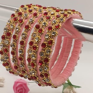 Könnte beinhalten: Ein Set aus rosafarbenen Armreifen mit goldfarbenen Akzenten und roten und klaren Edelsteinen. Die Armreifen sind gestapelt, was ihr aufwendiges Design zeigt. Eine rosa Rose und weiße Blumen befinden sich im Vordergrund.