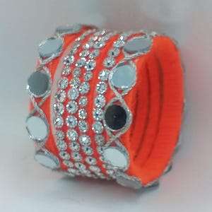 Könnte beinhalten: Ein Stapel orangefarbener Armreifen mit silbernen Spiegelakzenten und funkelnden Strasssteinen. Die Armreifen sind mit orangefarbenem Stoff umwickelt und weisen ein dekoratives Design aus Spiegeln und Strasssteinen auf.