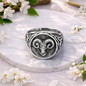 Może przedstawiać: Srebrny pierścionek z szczegółowym wzorem głowy barana pośrodku. Pierścionek ma ozdobną obwódkę i jest umieszczony na białej marmurowej powierzchni, otoczony białymi kwiatami. Pierścionek jest oznaczony "Handmade Mahi Silver Jewels 925 Silver".