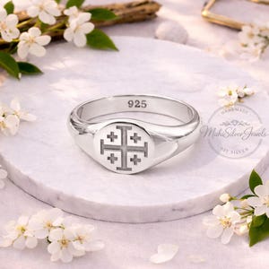 May include: A silver ring with a Jerusalem cross design. The ring is stamped with "925" and features a rounded face with the cross symbol. The ring is displayed on a white marble surface, surrounded by white flowers.