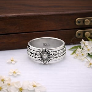 May include: A silver ring featuring a daisy flower design. The ring has a spinning band with a braided pattern. The flower's centre has a small, clear stone. The ring is marked with "925".