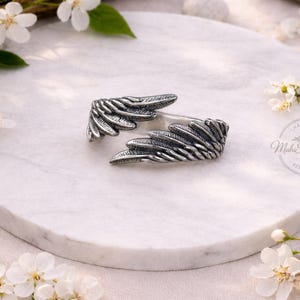 K&ouml;nnte beinhalten: Ein Silberring mit zwei detaillierten, &uuml;berlappenden Fl&uuml;gelmotiven und oxidierten Akzenten, die die Federstruktur hervorheben. Der Ring ist mit dem Text Mahi Silver Jewels 925 Silver gepr&auml;gt, was auf seine Zusammensetzung aus Sterlingsilber hinweist.