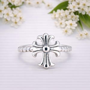 Puede incluir: Anillo plateado con un diseño de cruz detallado. La cruz tiene bordes curvos y ornamentados y un elemento circular central. La banda del anillo tiene un patrón texturizado y con cuentas. El fondo incluye flores blancas y una superficie blanca.