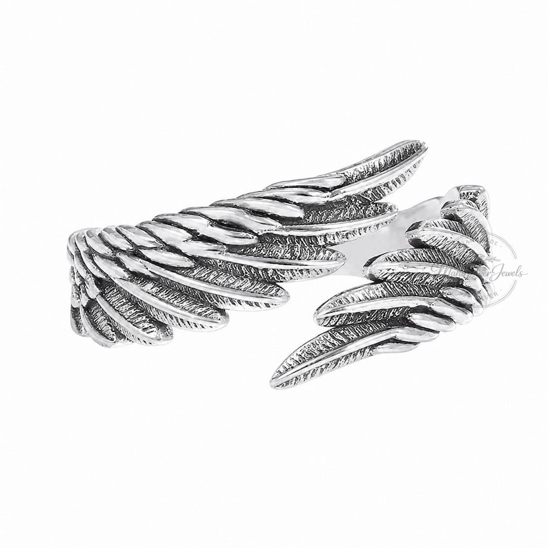 K&ouml;nnte beinhalten: Ein offener Silberring mit zwei detaillierten, &uuml;berlappenden Vogelfl&uuml;geln. Das Metall hat ein oxidiertes Finish, das die texturierten, geschichteten Federmuster auf der gesamten Oberfl&auml;che des Rings hervorhebt und f&uuml;r eine plastische Optik sorgt.